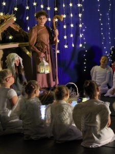 Na scenie przed szopka stoi uczeń odgrywający rolę Józefa. Chłopiec trzyma w jednej dłoni lampion a w drugiej pastorał. Przed nim na podłodze siedzą dziewczynki w strojach aniołów.