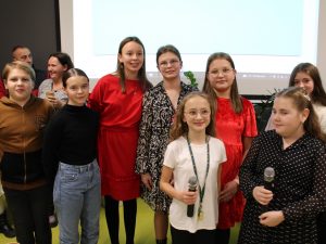 Uczniowie tworzący szkolny chórek pozują do wspólnego zdjęcia. Dwie uczennice stojące z przodu trzymają mikrofony.