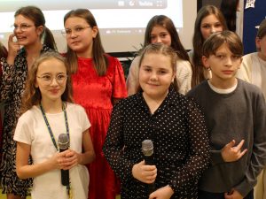 Zbliżenie na uczniów tworzących szkolny chórek. Dwie uczennice trzymają mikrofony. Obok nich stoi uczeń ze zgiętą w łokciu ręką i wyciągniętymi dwoma skrajnymi palcami u dłoni.