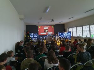 Na sali teatralnej siedzą uczniowie. Przed nimi widoczna jest świąteczna dekoracja ozdobiona lampkami. W tle na ekranie wyświetla się grafika na czerwonym tle z napisem "Jaka to melodia ?"
