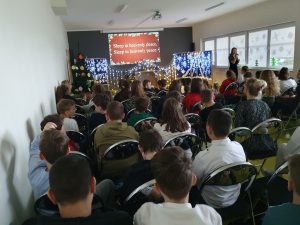 Uczniowie siedzą na sali teatralnej. Przed nimi widoczna jest dekoracja świąteczna ozdobiona lamkami. Za nią wyświetla się na ekranie tekst kolędy w języku angielskim.