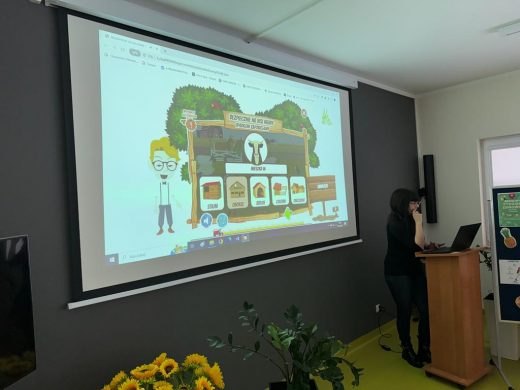 Zdjęcie wyświetlanej na rzutniku prezentacji oraz Pani stojącej za mównicą, na której ustawiony jest laptop.
