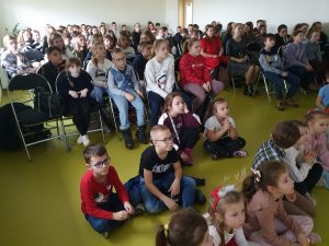 Uczniowie siedzą w sali teatralnej. Część z nich siedzi na podłodze, a za nimi starsi koledzy na krzesełkach. Sala wypełniona jest uczniami szkoły.