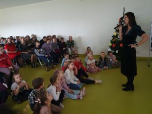 Pani Karolina stoi z mikrofonem na przeciwko dzieci. Najmłodsi uczniowie siedzą w rzędzie na podłodze, starsi za nimi na krzesełkach.