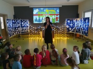 Pani Karolina stoi na środku sali teatralnej z mikrofonem. Przed nią na podłodze siedzą uczniowie. Za nią widoczna jest grafika z napisem KOLĘDY oraz dekoracja świąteczna ozdobiona lampkami.