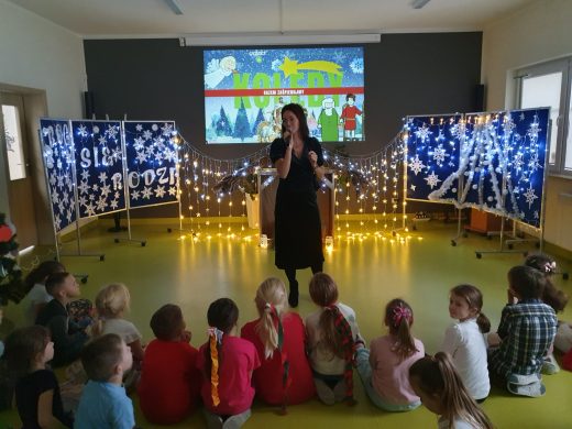 Pani Karolina stoi na środku sali teatralnej z mikrofonem. Przed nią na podłodze siedzą uczniowie. Za nią widoczna jest grafika z napisem KOLĘDY oraz dekoracja świąteczna ozdobiona lampkami.