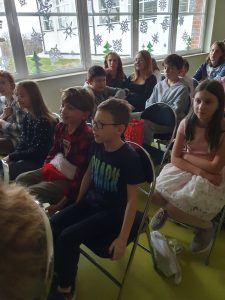 Uczniowie siedzą na sali teatralnej na krzesełkach i patrzą przed siebie.