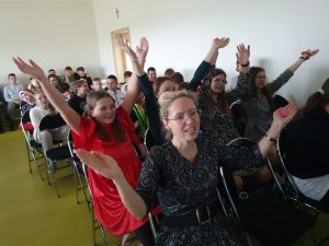 Uczniowie siedzą na sali teatralnej na krzesełkach. Na pierwszym planie widoczna jest nauczycielka oraz siedzące w rzędzie za nią uczennice z szeroko rozłożonymi do góry rękoma.
