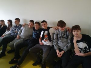 Uczniowie klasy siódmej siedzą w jednym rzędzie pod ścianą.