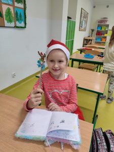Uczennica w czapeczce Mikołaja na głowie siedzi przy stoliku. Dziewczynka uśmiecha się patrząc na ołówek ozdobiony głową renifera, który trzyma w dłoni.