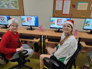 Dwie uczennice klasy szóstej pozują do wspólnego zdjęcia siedząc przy stanowiskach komputerowych w sali informatycznej. Na monitorach za dziewczynkami wyświetlają się tworzone przez nie postacie świętego Mikołaja.