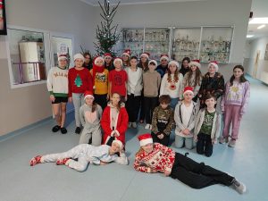 Uczniowie klasy szóstej pozują do grupowego zdjęcia na szkolnym korytarzu. Część dzieci na głowach ma czapki świętego Mikołaja i ubrana jest w piżamy.
