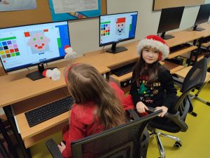 Uczennica w czapce Mikołaja pozuje do zdjęcia siedząc przy stanowisku komputerowym w sali informatycznej. Na monitorze za nią wyświetla się tworzona przez nią postać świętego Mikołaja. Obok widać pracującą przy komputerze inną uczennicę.