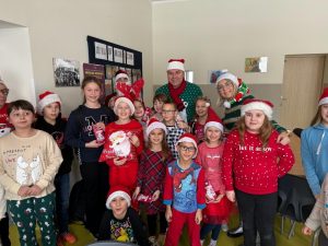 Uczniowie pozują w szkolnej stołówce do wspólnego zdjęcia z radnym gminy oraz jedną z nauczycielek. Część dzieci oraz dorośli mają na głowach czapki Mikołaja.