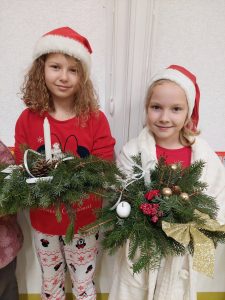 Dwie uczennice klasy drugiej w strojach mikołajkowych z czapkami na głowach trzymają w dłoniach przygotowane przez siebie stroiki świąteczne.
