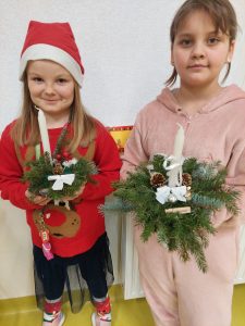 Dwie uczennice klasy drugiej trzymają w dłoniach przygotowane przez siebie stroiki świąteczne. Jedna z nich na głowie ma czapeczkę mikołajową.
