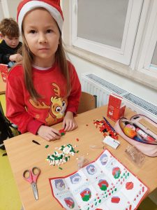 Uczennica klasy drugiej siedzi przy stoliku w czapeczce mikołajkowej. Przed nią leżą małe kawałki kolorowego papieru, wycinanka i przybory szkolne.