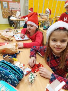 Dwie uczennice klasy drugiej podczas przygotowania świątecznych dekoracji. Dziewczynki mają na głowie świąteczne czapki mikołajkowe. Przed nimi na stolikach leżą przybory szkolne i wycinanki.