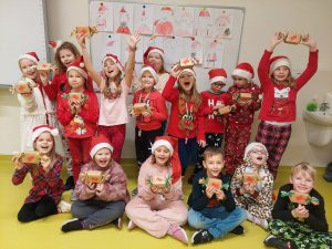 Uczniowie klasy drugiej pozują do wspólnego zdjęcia trzymając przed sobą otrzymane prezenty. Dzieci mają szeroko otwarte buzie. Część z nich stoi i wysoko w górę podnosi prezenty. Inni siedzą na podłodze i prezenty trzymają przed sobą.