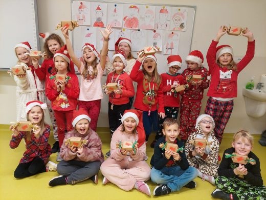 Uczniowie klasy drugiej pozują do wspólnego zdjęcia trzymając przed sobą otrzymane prezenty. Dzieci mają szeroko otwarte buzie. Część z nich stoi i wysoko w górę podnosi prezenty. Inni siedzą na podłodze i prezenty trzymają przed sobą.