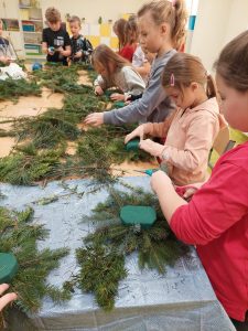 Uczniowie klasy drugiej stoją przy stole i wykonują stroiki świąteczne z gałązek iglaków.