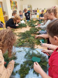 Uczniowie klasy drugiej stoją przy stole i wykonują stroiki świąteczne z gałązek iglaków.