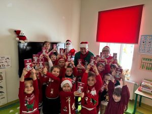 Uczniowie oddziały przedszkolnego pozują do wspólnego zdjęcia z wychowawczynią, świętym Mikołajem, radnym z gminy i Panią Dyrektor. Dzieci wyciągają wysoko w górę otrzymane czekolady.