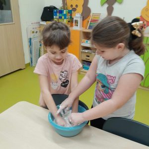 Dwie uczennice oddziału przedszkolnego wkładają swoje dłonie do miski z masą solną. Dziewczynki patrzą na nie.