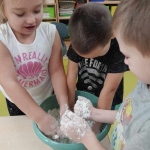 Troje uczniów oddziału przedszkolnego wkłada swoje dłonie do miski z masą solną. Chłopiec stojący po prawej dłoni ogląda swoje dłonie oblepione masą solną.