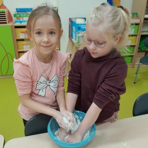 Dwie uczennice oddziału przedszkolnego wkładają swoje dłonie do miski z masą solną. Jedna z dziewcząt trzyma w dłoniach kulkę z masy solnej i patrzy się przed siebie.