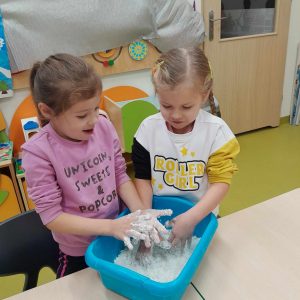 Dwie uczennice oddziału przedszkolnego wkładają swoje dłonie do miski z masą solną. Dziewczynki patrzą na nie.
