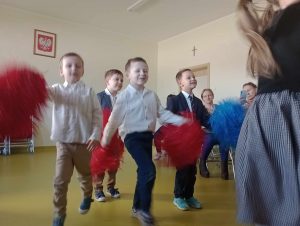 Uczniowie oddziału przedszkolnego tańczą z czerwonymi i niebieskimi pomponami.