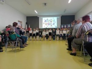 Uczniowie oddziału przedszkolnego stoją w jednym rzędzie. Dzieci ubrane są w stroje galowe. W tle wyświetla się na rzutniku kolorowy napis "kochamy was". Na przeciwko na krzesełkach siedzą babcie i dziadkowie.