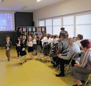 Uczniowie z oddziału przedszkolnego stoją w grupce trzymając w dłoniach upominki dla swoich dziadków, którzy siedzą na krzesełkach.