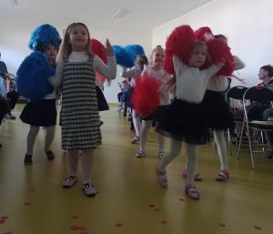 Dziewczynki z oddziału przedszkolnego tańczą trzymając w dłoniach niebieskie i czerwone pompony.