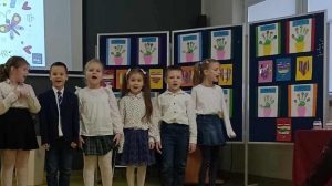 Uczniowie oddziału przedszkolnego stoją w jednym rzędzie. Dzieci ubrane są na galowo i mają szeroko otwarte buzie. Za nimi na tablicy przyczepione są laurki wykonane dla babć i dziadków.