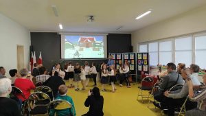 Uczniowie oddziału przedszkolnego tańczą w parach na sali teatralnej. Dzieci obserwują babcie i dziadkowie siedzące na przeciwko nich na krzesełkach. W tle na rzutniku wyświetla się obrazek przedstawiający zimowy krajobraz i uśmiechniętą dziewczynkę stojącą na jego tle.