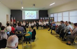 Uczniowie oddziału przedszkolnego stoją w jednym rzędzie. Dzieci ubrane są w stroje galowe. W tle wyświetla się na rzutniku kolorowy napis "kochamy was". Na przeciwko na krzesełkach siedzą babcie i dziadkowie.