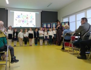 Uczniowie oddziału przedszkolnego stoją w jednym rzędzie obok swojej wychowawczyni. Pani trzyma w dłoni mikrofon, a dzieci ubrane są w stroje galowe. W tle wyświetla się na rzutniku kolorowy napis "kochamy was". Na przeciwko na krzesełkach siedzą babcie i dziadkowie.