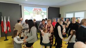 Dziadkowie wraz ze swoimi wnuczętami tańczą na sali teatralnej w parach. W tle wyświetla się na rzutniku obrazek z zimowym krajobrazem.