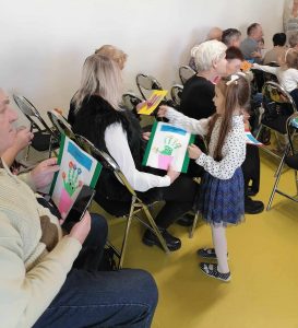 Uczennica oddziału przedszkolnego daje siedzącej przed nią babci laurkę. Obok widać innych dziadków trzymających swoje laurki.