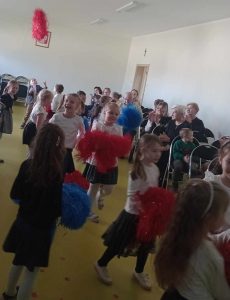 Dziewczynki z oddziału przedszkolnego tańczą trzymając w dłoniach niebieskie i czerwone pompony. Jedna z uczennic rzuciła w górę swój czerwony pompon.