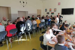 Babcie i dziadkowie siedzą przy długim stole na szkolnej stołówce. W drugim rzędzie przy stole siedzą uczniowie oddziału przedszkolnego. Na stole ustawione są słodkie przekąski i talerzyki.