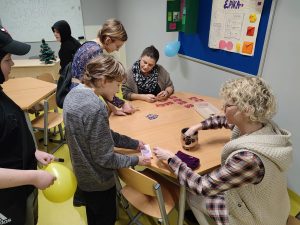 Przy stoliku siedzą dwie nauczycielki. Jedna z nich wkłada rękę w srebrny pojemnik. Obok stoi uczeń trzymający w dłoni banknot. Na przeciwko siedząca nauczycielka spogląda na kupon uczennicy stojącej obok niej.