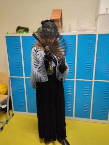 Uczennica pozuje w swoim przebraniu na tle kolorowych szafek. Dziewczyna ubrana jest w czarną długą suknię i wydzierganą szarą pelerynę. Twarz zakrywa koronkowym, czarnym wachlarzem.