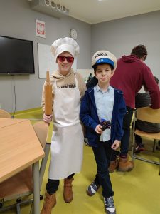 Dwoje uczniów pozuje do wspólnego zdjęcia. Jeden z nich przebrany jest za kucharza. Na oczach ma przeciwsłoneczne okulary a w dłoni trzyma wałek do ciasta. Drugi chłopiec przebrany jest za policjanta i trzyma w dłoni zabawkowy pistolet.