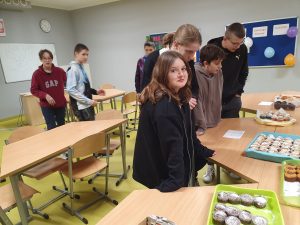 Czworo uczniów stoi w rzędzie przy stołach na których ustawione są tace z babeczkami. W tle widać troje innych uczniów. Uczennica widoczna na pierwszym planie patrzy w stronę obiektywu.