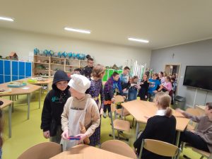 Uczniowie stoją w kolejce w jednej z klas. Na pierwszym planie widoczny jest chłopiec w stroju kucharza trzymający wałek do ciasta. Za nim stoi chłopiec w czarnej bluzie patrzący na niego.