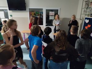 Uczniowie zgromadzeni są na szkolnym korytarzu. Na przeciwko nich stoi z mikrofonem nauczycielka.