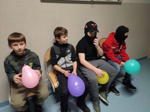 Czterech uczniów siedzi na krzesełkach na szkolnym holu obok siebie. Każdy z nich trzyma balon w innym kolorze. Dwóch chłopców ma na głowie kominiarki.